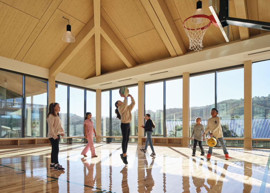 Interior shot of Ohana Montage Health Facility, inside the recreational basketball court. The court features an exposed timber roof, wood floors, and floor to ceiling windows. Children are shown playing a game of basketball.