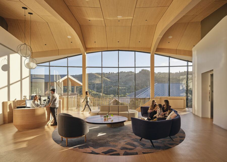 Interior shot of the Ohana Montage Health Facility, with a central lobby area featuring exposed wood ceilings facing towards the large window, showing people sitting on chairs and milling about.